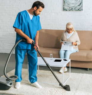 caregiver vacuuming the floor while senior woman is sitting on the sofa